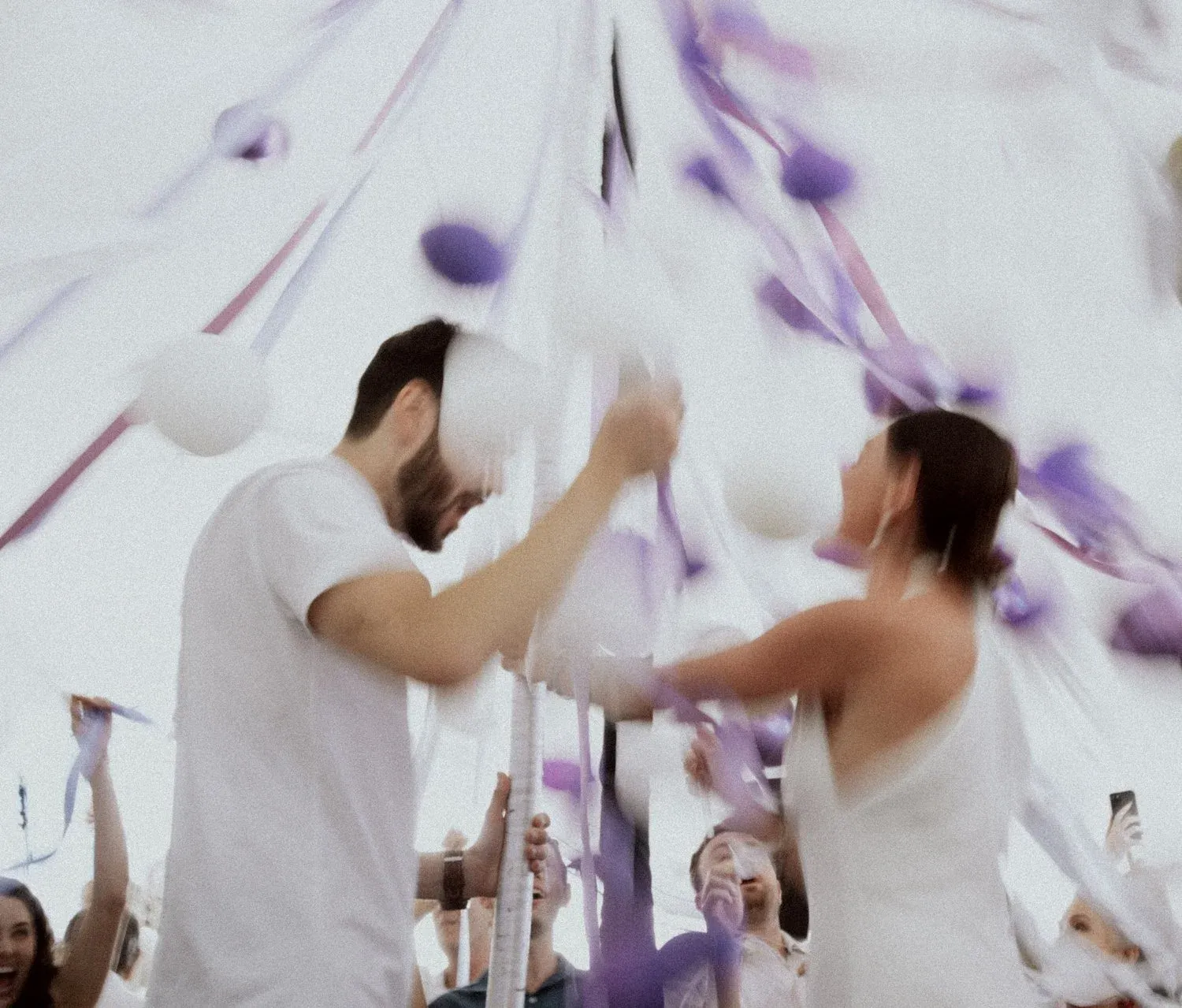 A man and a woman holding a large white and lavender umbrella at an outdoor event with other people around, some taking photos.