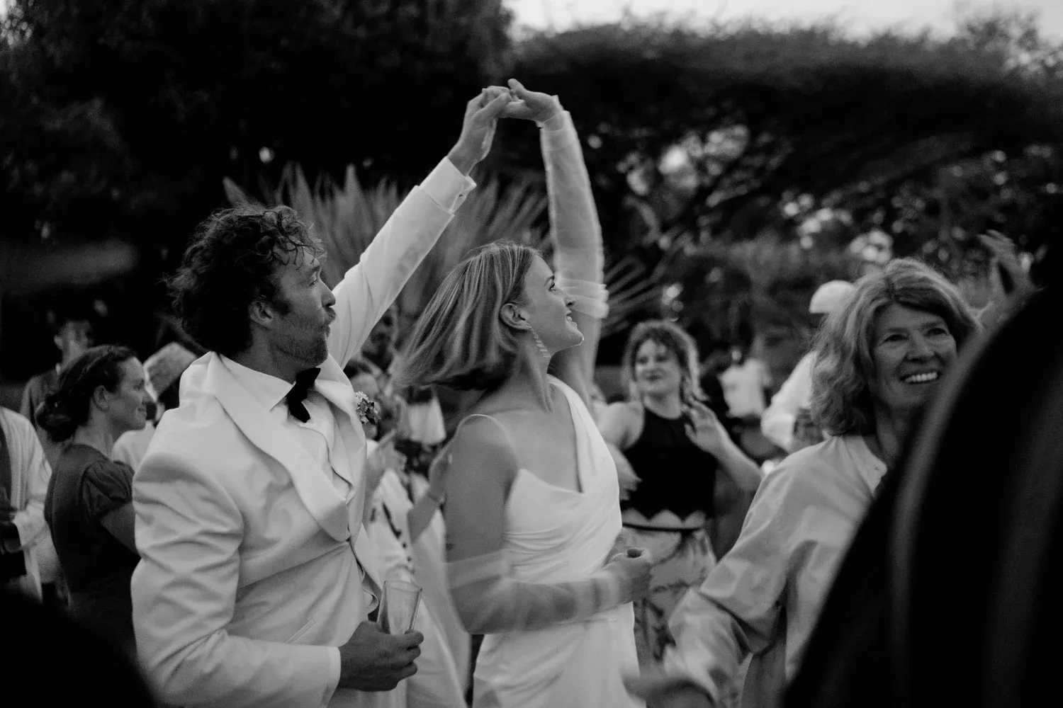 A black and white photo of a wedding reception with a couple dancing, the man in a white tuxedo holding the woman's hand, surrounded by guests smiling and dancing outdoors.