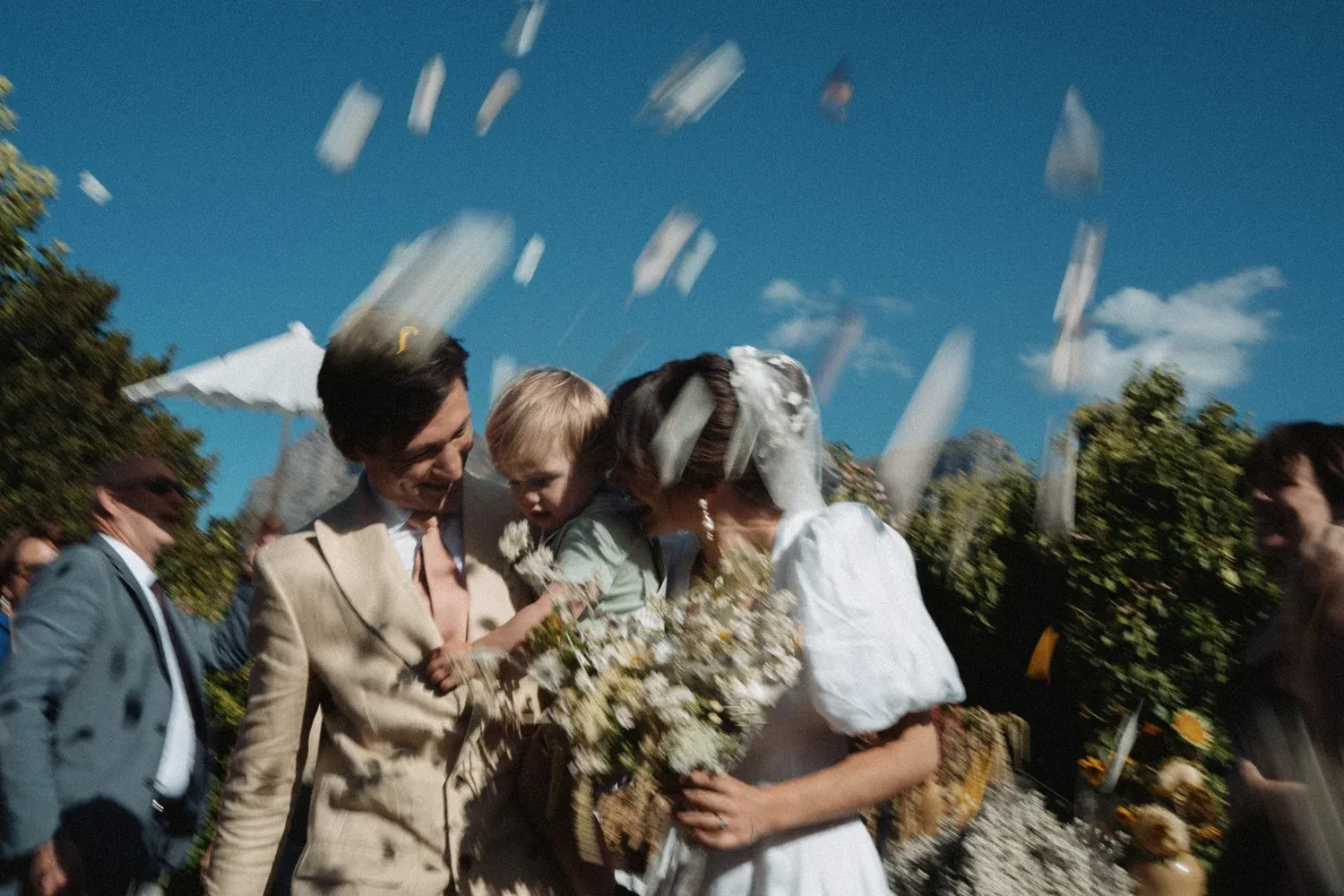 A wedding celebration outdoors with confetti in the air, a bride holding a bouquet, a groom smiling, and a young boy with guests in the background.