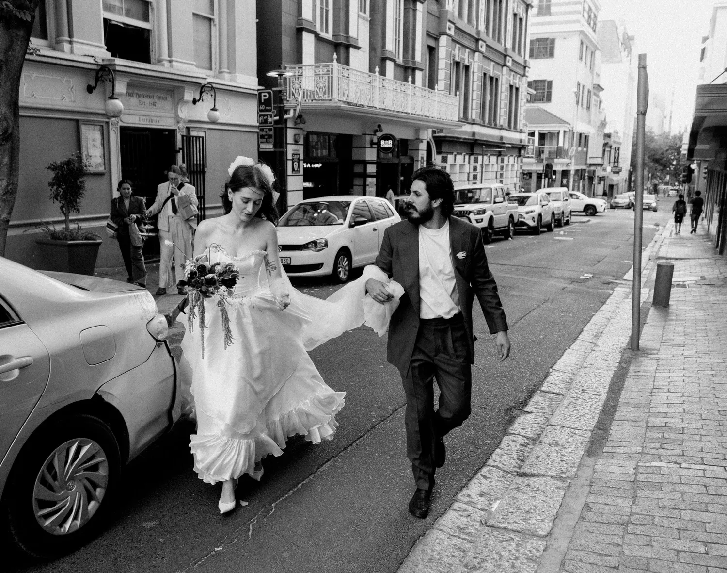 A bride in a wedding dress and a groom in a suit walking on a city street, with the bride holding a bouquet of flowers and the groom guiding her as they walk past parked cars and pedestrians.