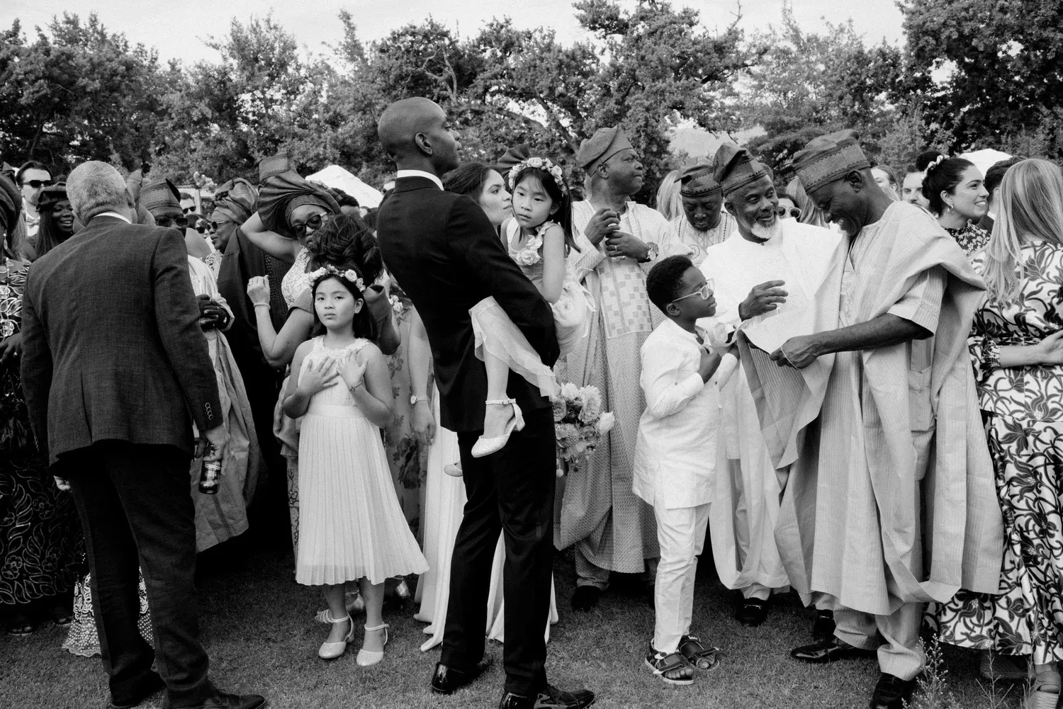 Group of people gathered outdoors for a celebration or wedding, with diverse individuals in formal attire, some children, and trees in the background.