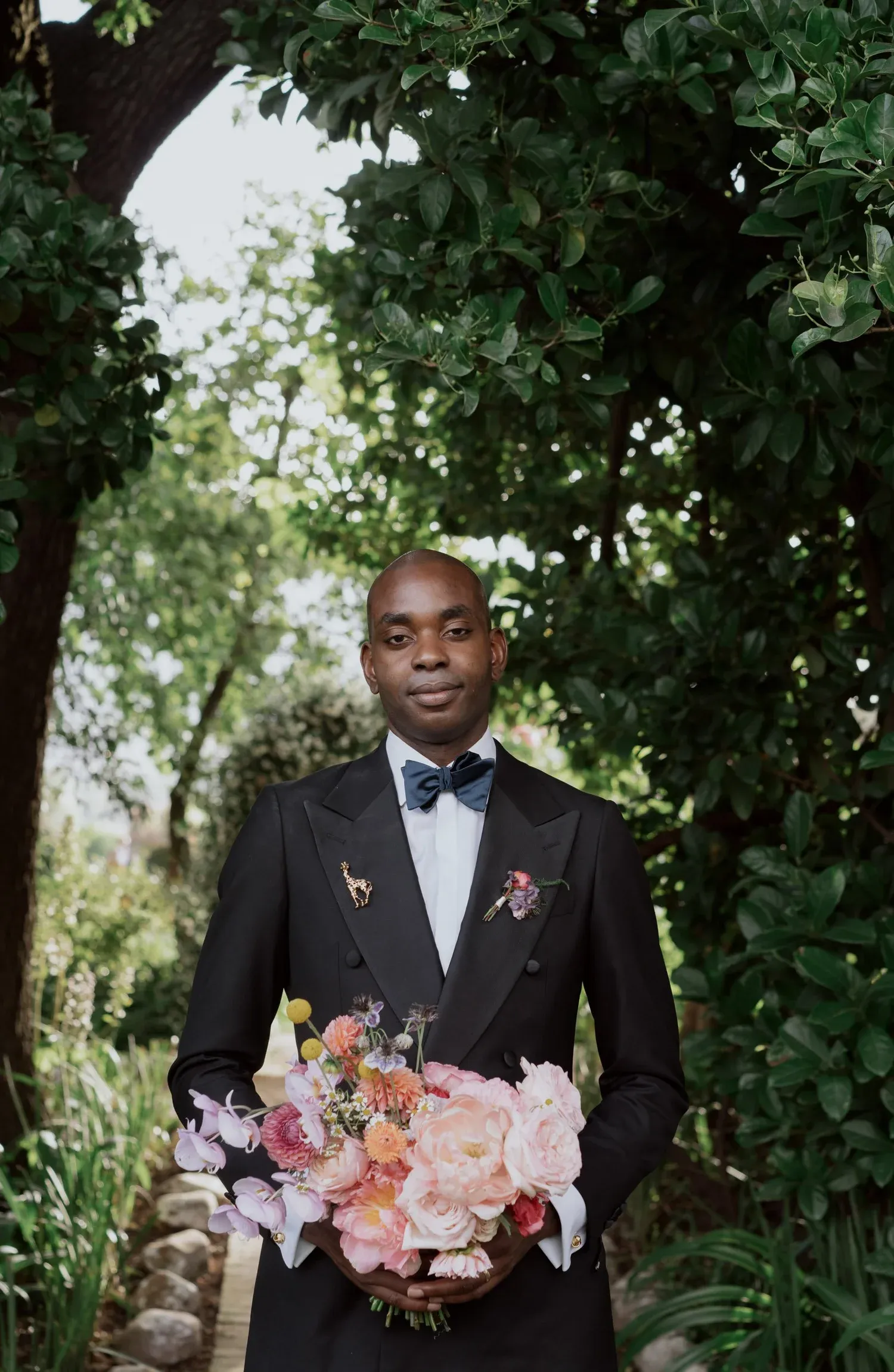 A man dressed in a formal tuxedo with a bow tie, holding a bouquet of pink and purple flowers, standing outdoors among trees.