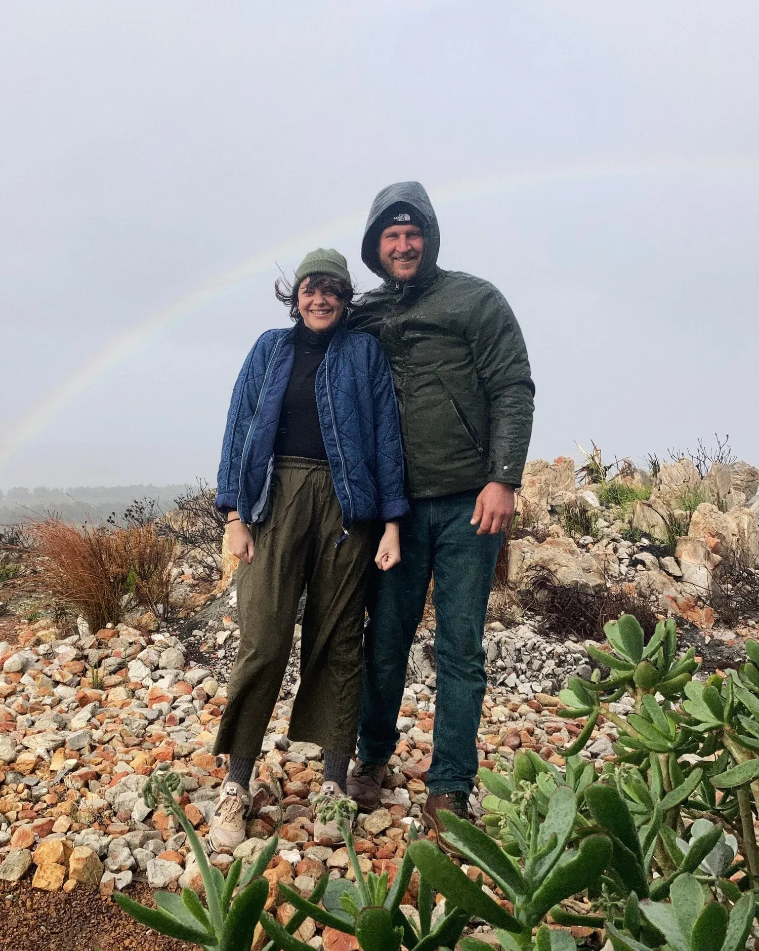 A man and a woman standing on rocky terrain outdoors, smiling, with a rainbow in the background during a rainy day.